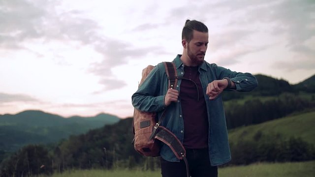 Young man tourist traveller with backpack hiking in nature, checking the time.