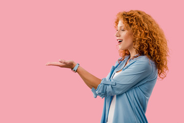 excited redhead woman presenting something isolated on pink