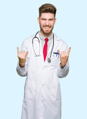 Fototapeta premium Young handsome doctor man wearing medical coat shouting with crazy expression doing rock symbol with hands up. Music star. Heavy concept.