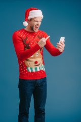 angry man in santa hat shouting and showing middle finger to smartphone, isolated on blue