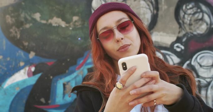 Close up of the young pretty woman in hipster style, with red hair, in sunglasses and hat typing and texting a message on the smartphone while standing on the graffity background in sunlight. Outside.