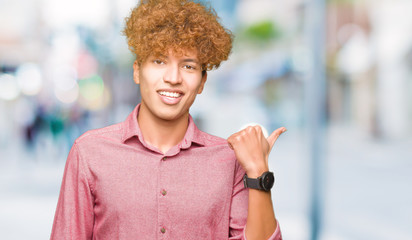 Young handsome business man with afro hair smiling with happy face looking and pointing to the side with thumb up.