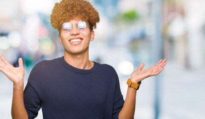 Young handsome man with afro wearing glasses clueless and confused expression with arms and hands...