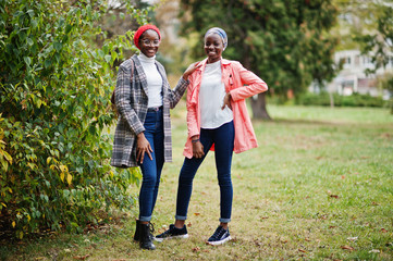 Two young modern fashionable, attractive, tall and slim african muslim womans in hijab or turban head scarf and coat posed at park together.