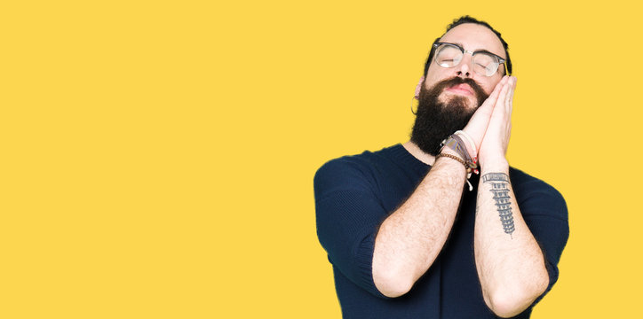 Young hipster man with long hair and beard wearing glasses sleeping tired dreaming and posing with hands together while smiling with closed eyes.