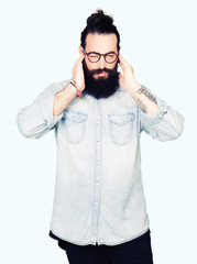 Fototapeta premium Young hipster man with long hair and beard wearing glasses covering ears with fingers with annoyed expression for the noise of loud music. Deaf concept.