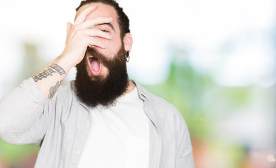 Young man with long hair, beard and earrings peeking in shock covering face and eyes with hand, looking through fingers with embarrassed expression.