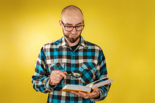 Research and business. A bald man with a beard and glasses, holding a book in his hands and carefully studying it through a magnifying glass. Yellow background. Copy space