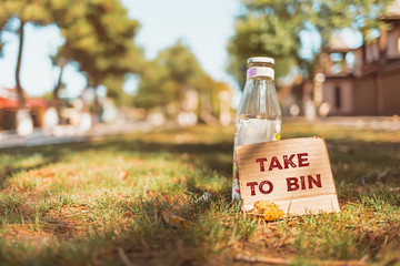 The concept of environmental pollution and waste sorting. In the backyard, in the sun, there is a glass bottle in the grass, with a cardboard, with the text TAKE TO BIN. Copy space