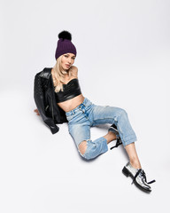 Biker punk woman in torn jeans, leather top and jacket, winter hat and brutal shoes sitting on white looks up in camera