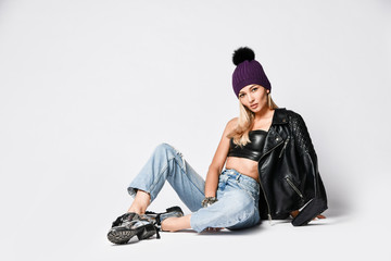 Biker style woman in torn jeans, leather top and jacket, winter hat and brutal shoes sitting on white looks in camera