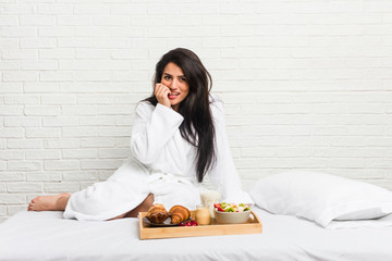 Young curvy woman taking a breakfast on the bed biting fingernails, nervous and very anxious.