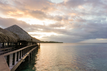 A wooden walkway with thatched roof bungalows leads out into the lagoon on the island of Moorea in French Polynesia at sunset; ocpy space