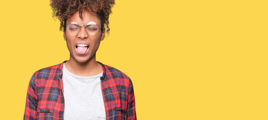 Beautiful young african american woman wearing glasses over isolated background sticking tongue out...