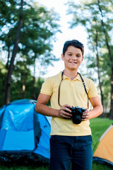 happy boy holding digital camera and smiling near camps in park