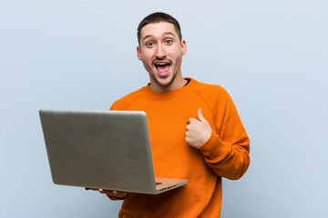 Young caucasian man holding a laptop surprised pointing at himself, smiling broadly.