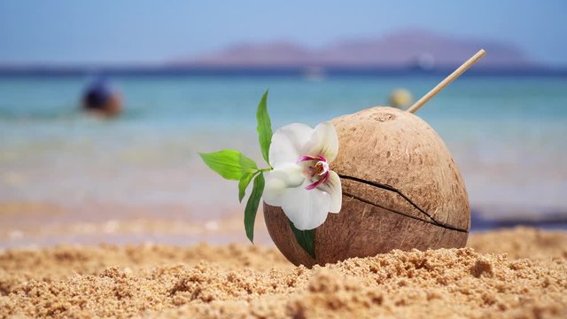 Coco cocktail in a coconut stands in the sand on an ocean beach. Small waves rise behind. Female legs in defocus pass by.