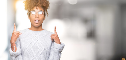 Beautiful young african american woman wearing winter sweater over isolated background amazed and surprised looking up and pointing with fingers and raised arms.