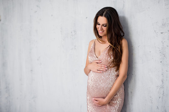 happy beautiful pregnant woman in shiny evening dress looking at her tummy over gray wall background with copy space