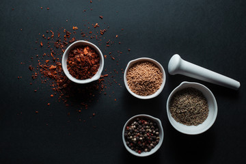 Paprika, mustard, caraway seeds and a mixture of peppercorns in white ceramic bowls next to the mortar isolated on black background