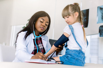 Little Caucasian girl at paediatrician office measuring blood pressure. Young African woman doctor...