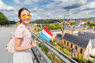 A happy Asian traveller girl holds the flag of Luxembourg and admires the Grund area from the observation deck. Tourism, recreation and life in the country.