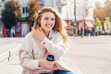 Girl student sitting on the steps with a cup of coffee and laughing out loud. Girl walks around the city and laughs