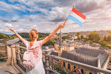 A happy traveller girl holds the flag of Luxembourg and admires the Grund area from the observation deck. Tourism, recreation and life in the country. - obrazy, fototapety, plakaty