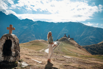 Outdoor travel lifestyle smiling woman tourist posing against the backdrop of the mountains and medieval church / monastery. Safari style girl with hat