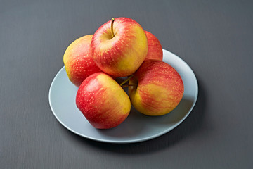 Heap of red fresh whole ripe apples full of vitamins lies on circle ceramic plate and old dark scratched concrete table on kitchen. Harvest or diet concept. Close-up