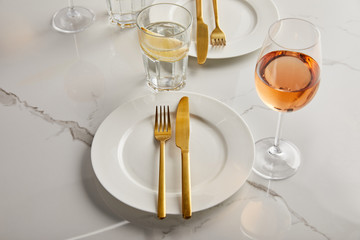 golden knives and forks on white plates near glasses with rose wine and lemon water on marble table