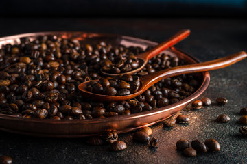Round copper tray, two wooden spoons full of kopi luwak coffee beans on dark background, closeup view