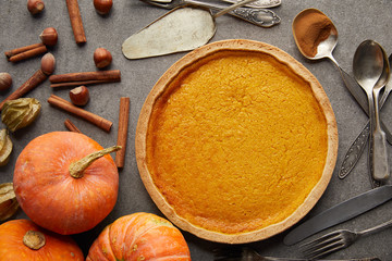 top view of delicious pumpkin pie near whole pumpkins, cutlery, hazelnuts and cinnamon sticks on grey stone surface