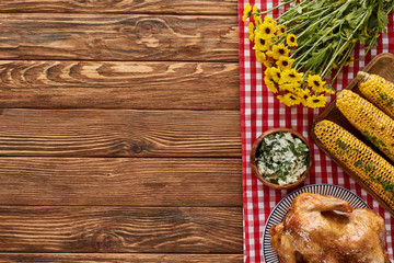 Fototapeta na wymiar top view of roasted turkey, grilled corn and yellow wildflowers on red checkered napkin on wooden table