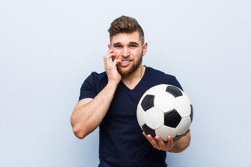 Young caucasian man holding a soccer ball biting fingernails, nervous and very anxious.