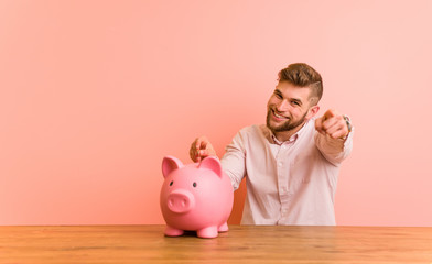 Young caucasian man sitting with a piggy bank cheerful smiles pointing to front.