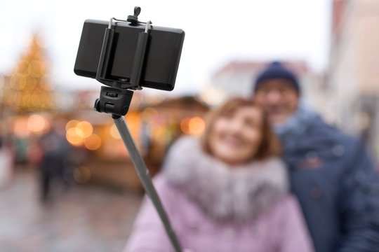technology, winter holidays and gadgets concept - close up of smartphone on selfie stick taking picture of senior couple at christmas market on town hall square in tallinn, estonia