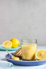A jar of lemon curd or custard with a spoon and cookies on a blue plate and fresh lemon. Selective focus, copy space.