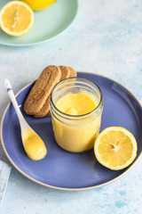 A jar of lemon curd or custard with a spoon and cookies on a blue plate and fresh lemon. Selective focus, copy space.