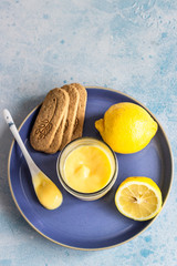 A jar of lemon curd or custard with a spoon and cookies on a blue plate and fresh lemon. Selective focus, copy space.