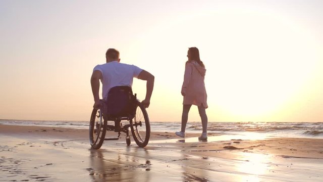 Handicapped man in wheelchair and his girlfriend on a beach at sunset