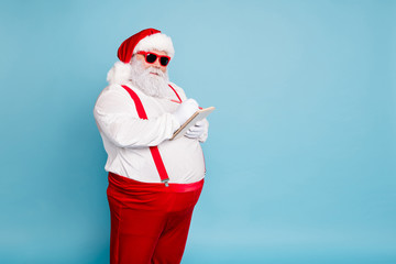 Turned photo of funny funky fat santa claus in modern eyewear with big belly abdomen write with pencil in note book make wish list wear red suspenders isolated over blue color background