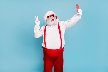 Fototapeta na wymiar Ho-ho-ho. Portrait of overweight style funny hipster santa claus blogger in cap hat take selfie on cellphone make v-signs enjoy christmas wear red trendy suspenders isolated blue color background