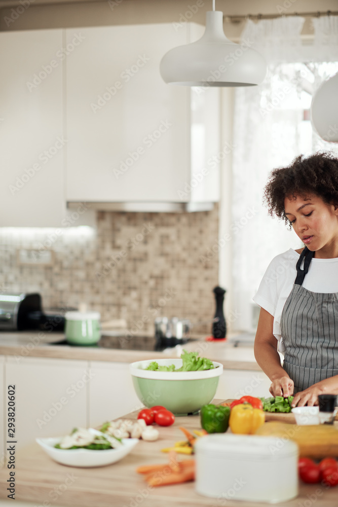 Wall mural Attractive mixed race woman in apron cutting lettuce for salad. Kitchen interior.