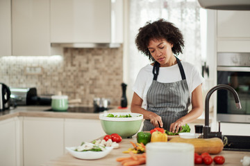 Attractive mixed race woman in apron cutting lettuce for salad. Kitchen interior.