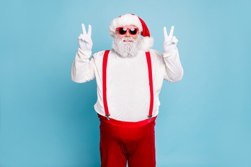 Fototapeta na wymiar Portrait of funny funky fat overweight santa in modern eyeglasses with big abdomen belly make v-signs enjoy christmas time wear suspenders white overalls isolated over blue color background