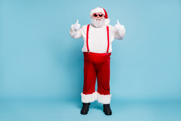 Full body photo of funny funky fat santa claus with big belly abdomen in eyewear show thumb up recommend winter season shopping ads wear red overalls boots pants isolated over blue color background © deagreez