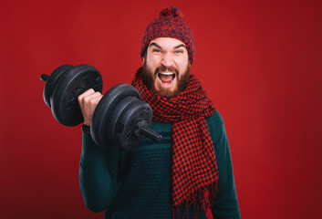 Strong bearded man, wearing winter clothes is lifting up a black dumbbell. Go fit and healthy, holiday offer on red background.