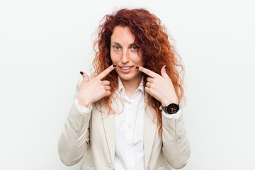 Fototapeta premium Young natural redhead business woman isolated against white background smiles, pointing fingers at mouth.
