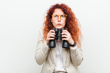 Obraz premium Young caucasian business redhead woman holding a binoculars biting fingernails, nervous and very anxious.